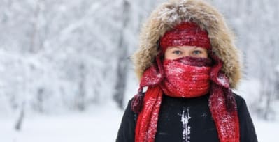 woman in snow wearing: winter coat, knit hat with long ear flaps, hood pulled up, and thick knit scarf wrapped around her neck and the lower section of her face. Only her eyes are visible.